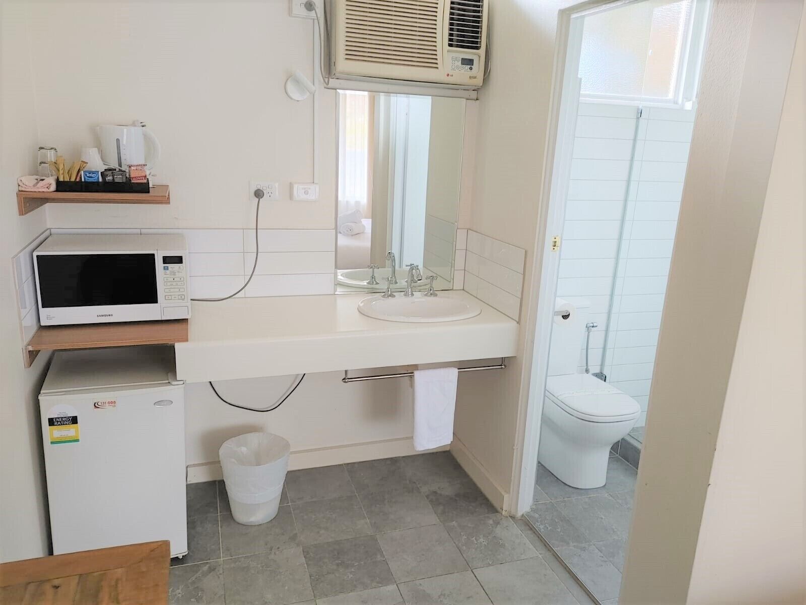 Un baño compacto con un lavamanos blanco, un microondas, un refrigerador pequeño, un cesto de basura y un inodoro.
