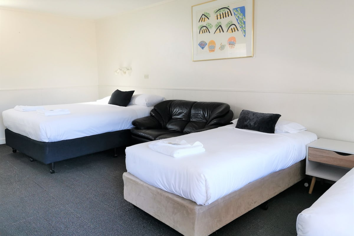 A hotel room with two beds, a black leather sofa, a wooden nightstand, and a framed picture on the wall.