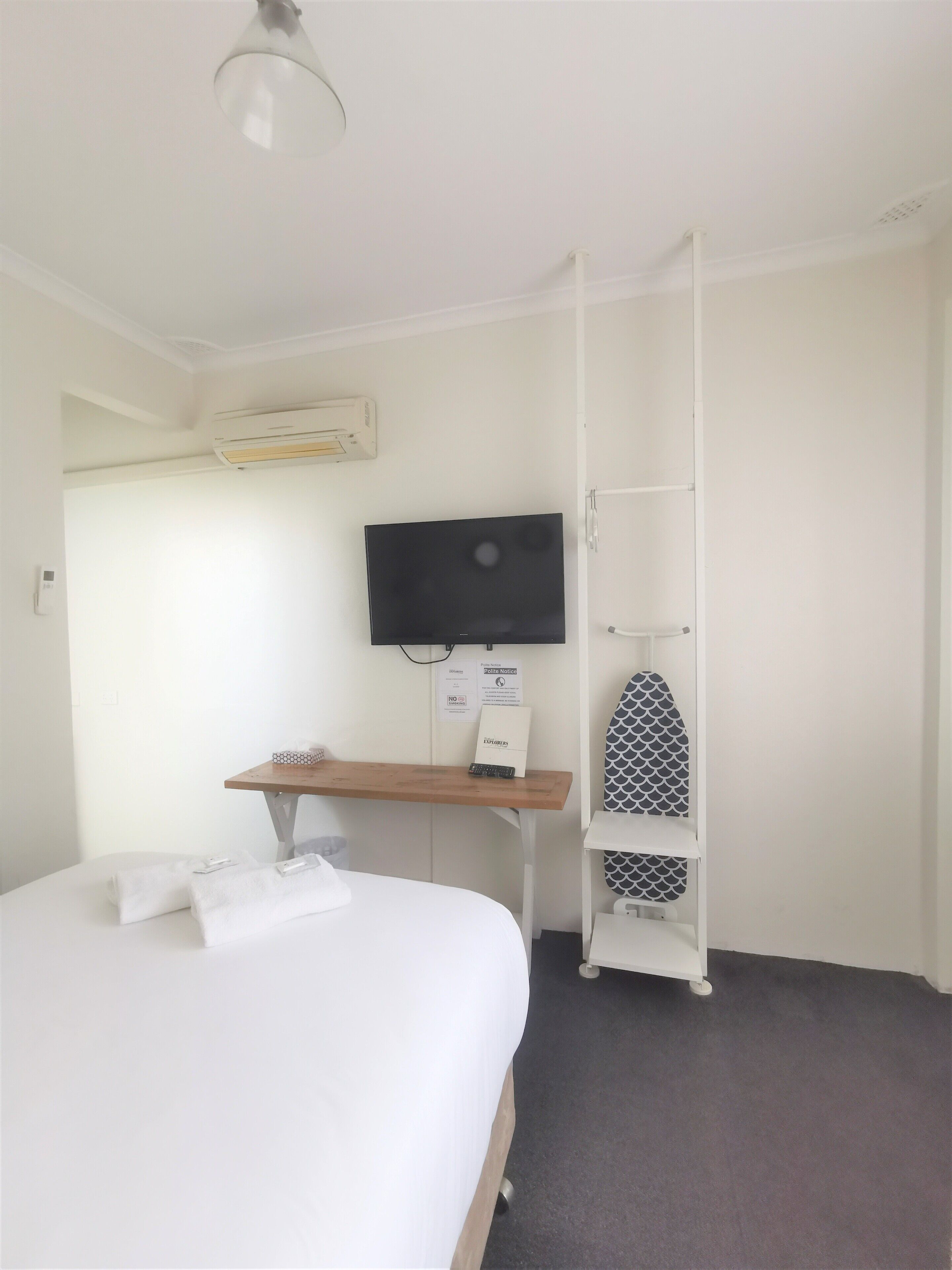 A hotel room with a bed, a desk with a flat-screen TV, an ironing board, and a wall-mounted air conditioner.