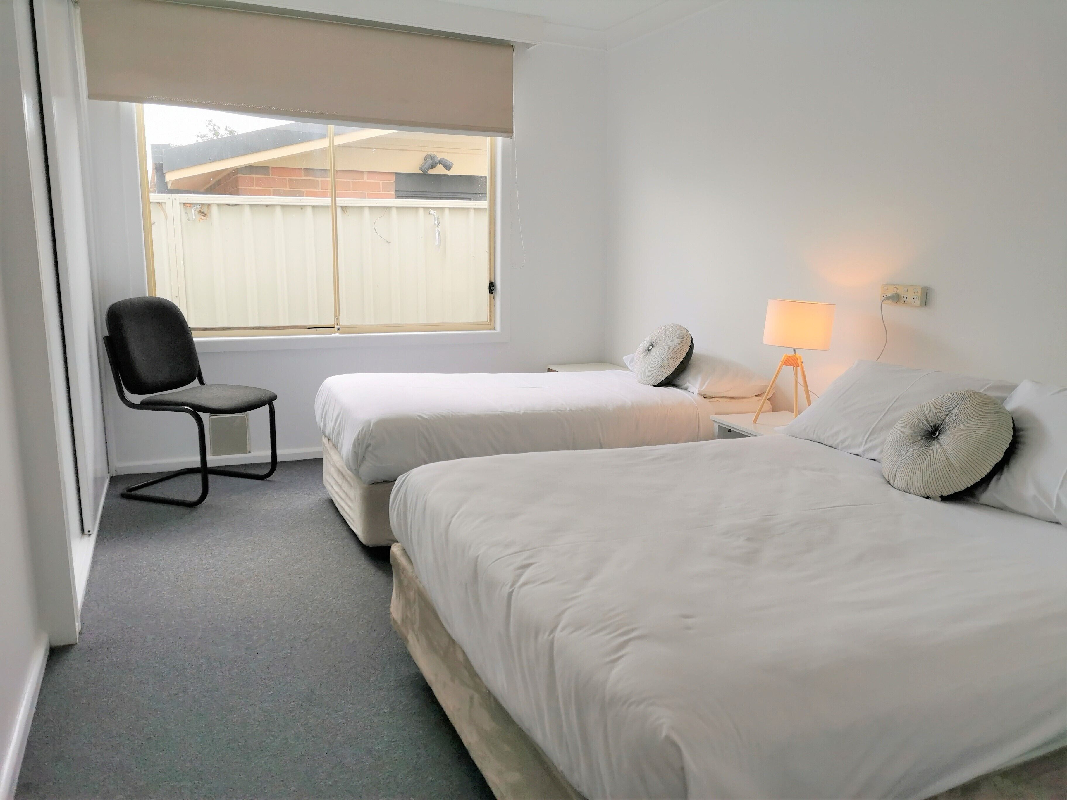 A hotel room with two beds, a chair, a lamp, and a window with a view of a fence.
