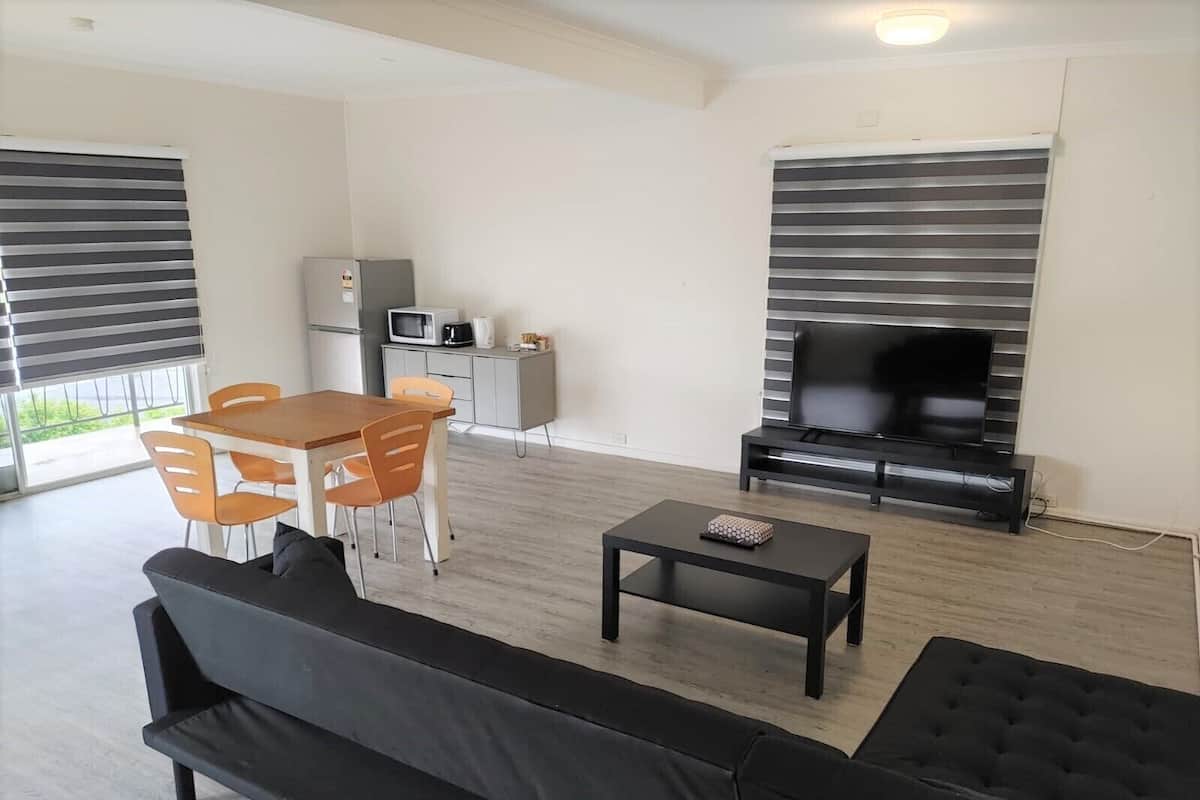 A modern living room with a sofa, a dining table with chairs, a TV, and a kitchen area with appliances.