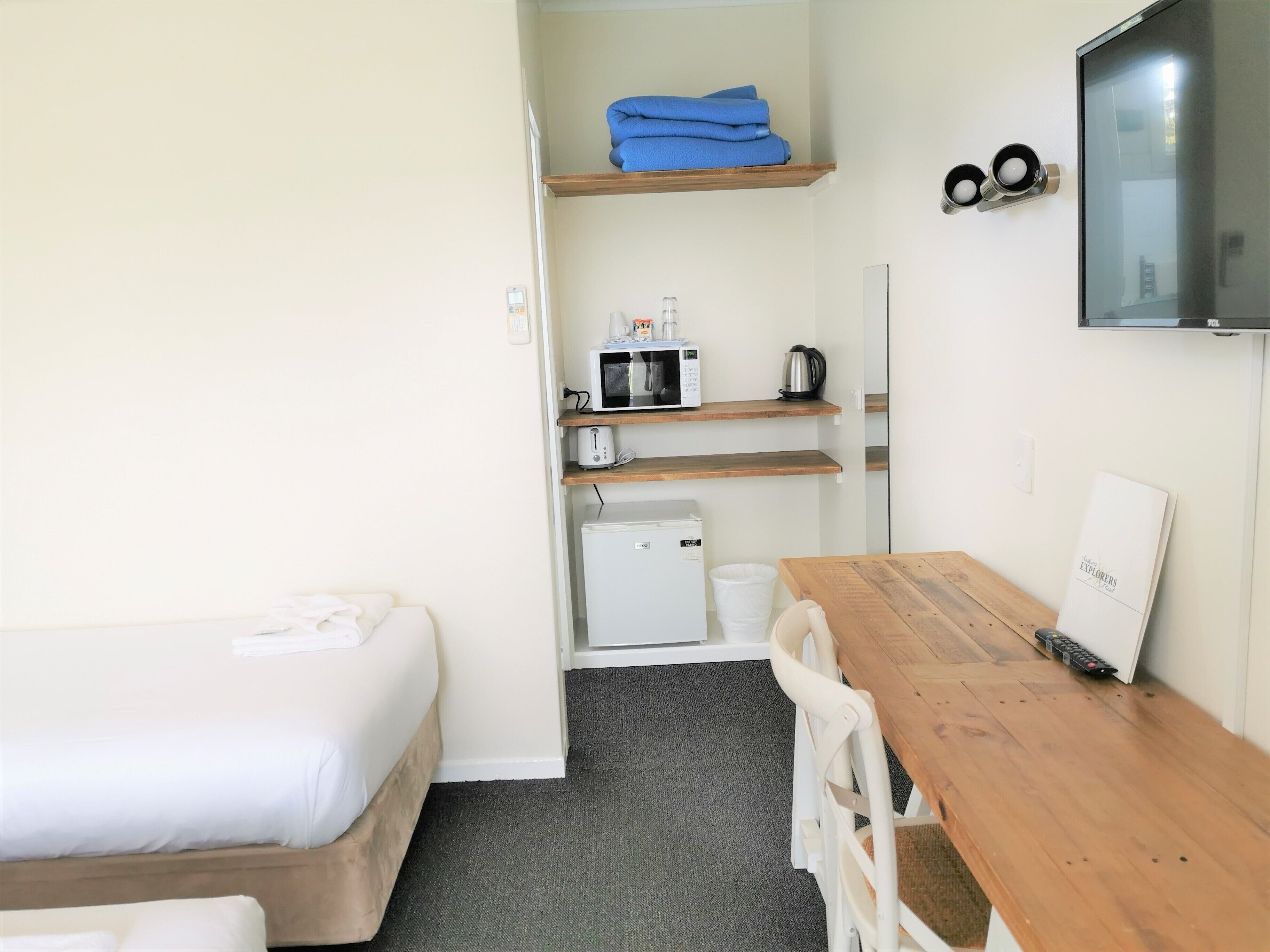Habitación de hotel compacta con una cama individual, un escritorio de madera, televisor y una kitchenette con microondas, tostadora y hervidor.