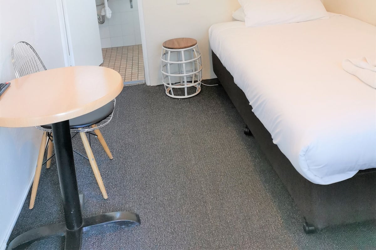 A hotel room with a bed, a small round table with a chair, a mirror, and a white air conditioning unit on the wall.