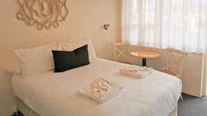 A neatly made bed with white linens and a black pillow, a small round table with a chair, and a window with sheer curtains. - Bathurst Explorers Motel (Bathurst)