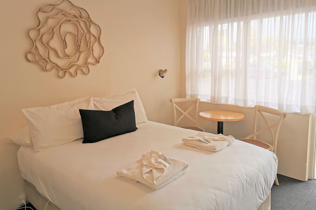 A neatly made bed with white linens and a black pillow, a small round table with a chair, and a window with sheer curtains.