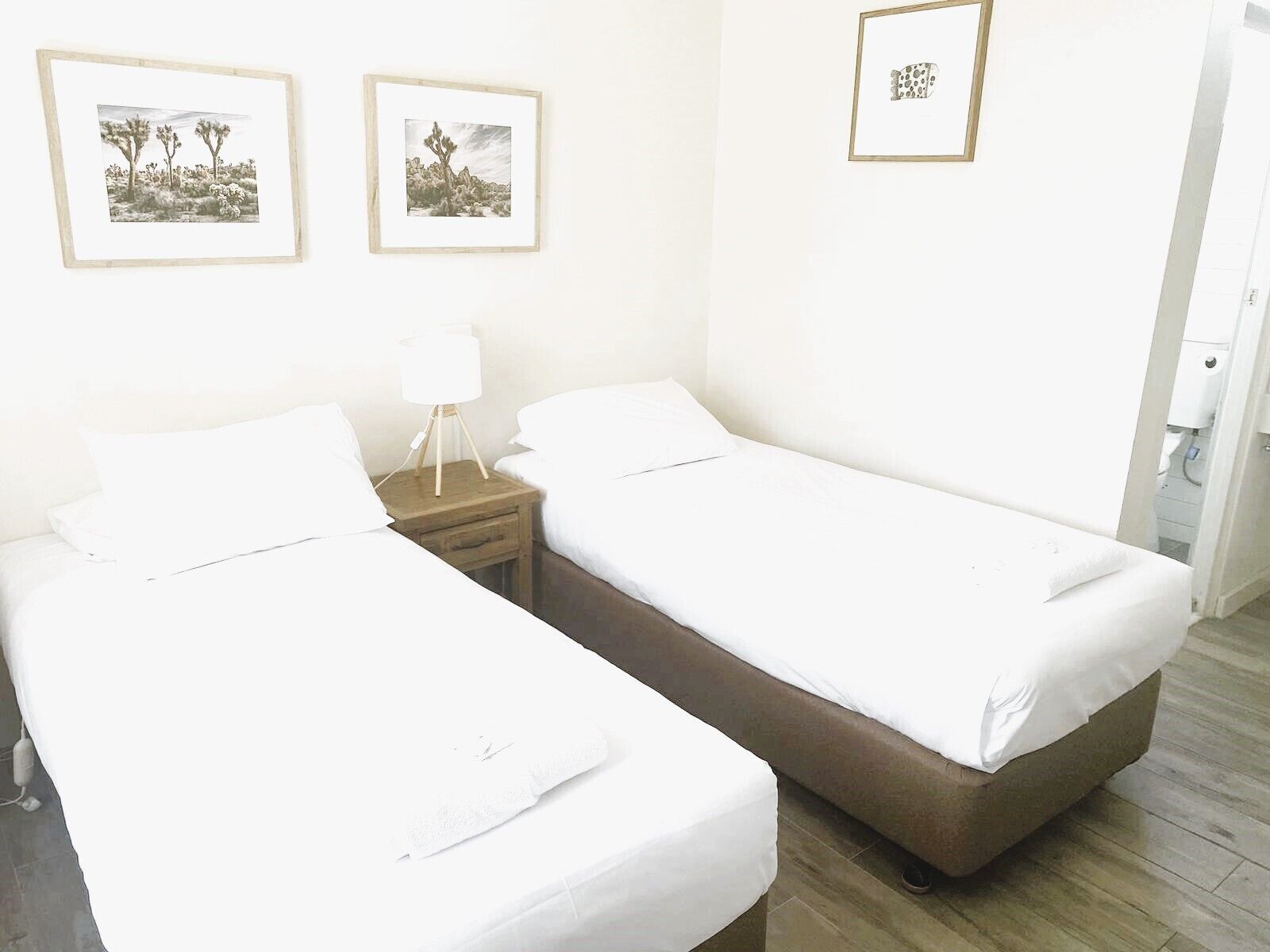 Habitación de hotel con dos camas individuales, una mesita de noche con lámpara y dos cuadros en la pared.