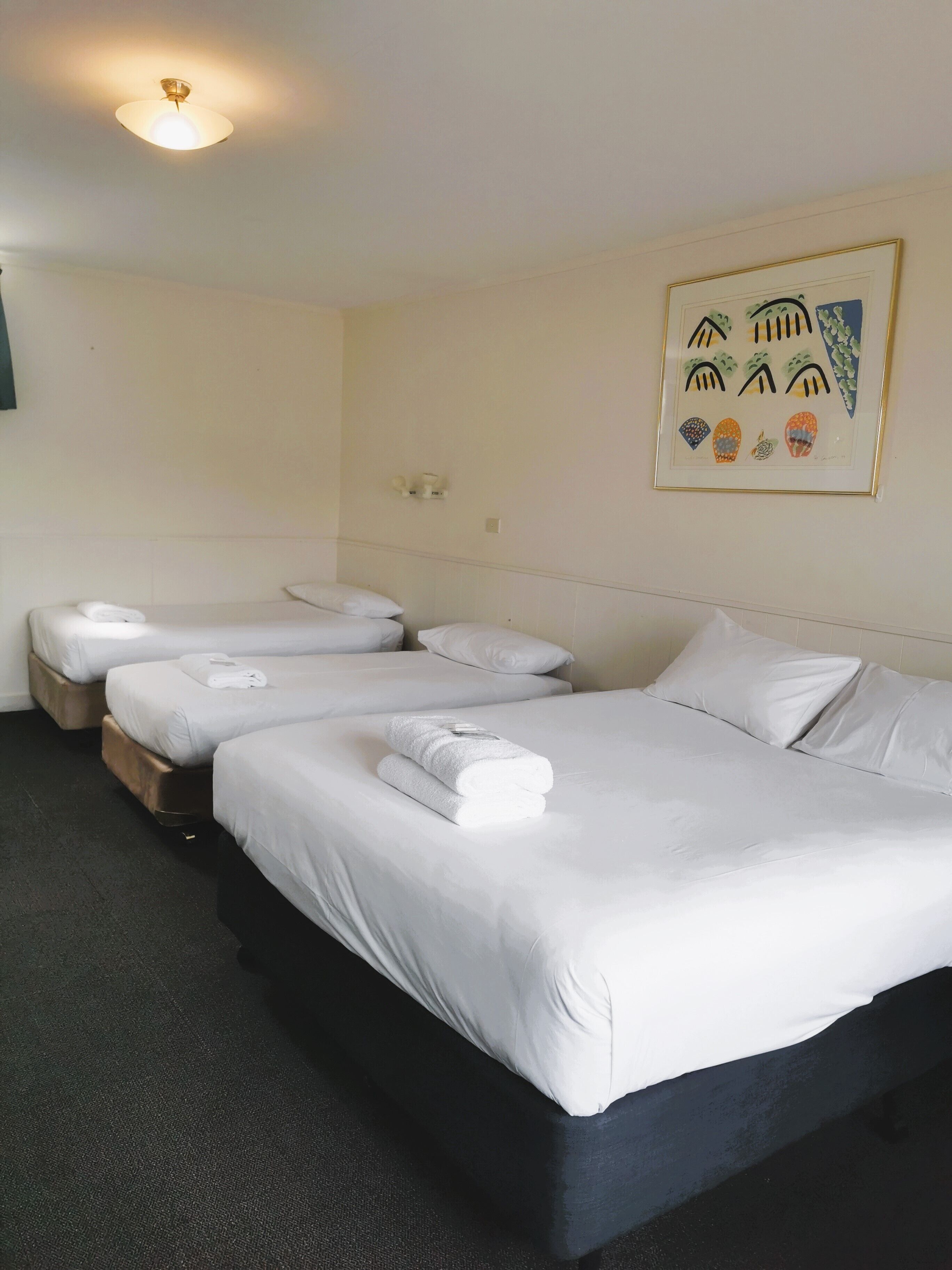A hotel room with three single beds, each with white linens and towels, a dark carpeted floor, and a ceiling light fixture.