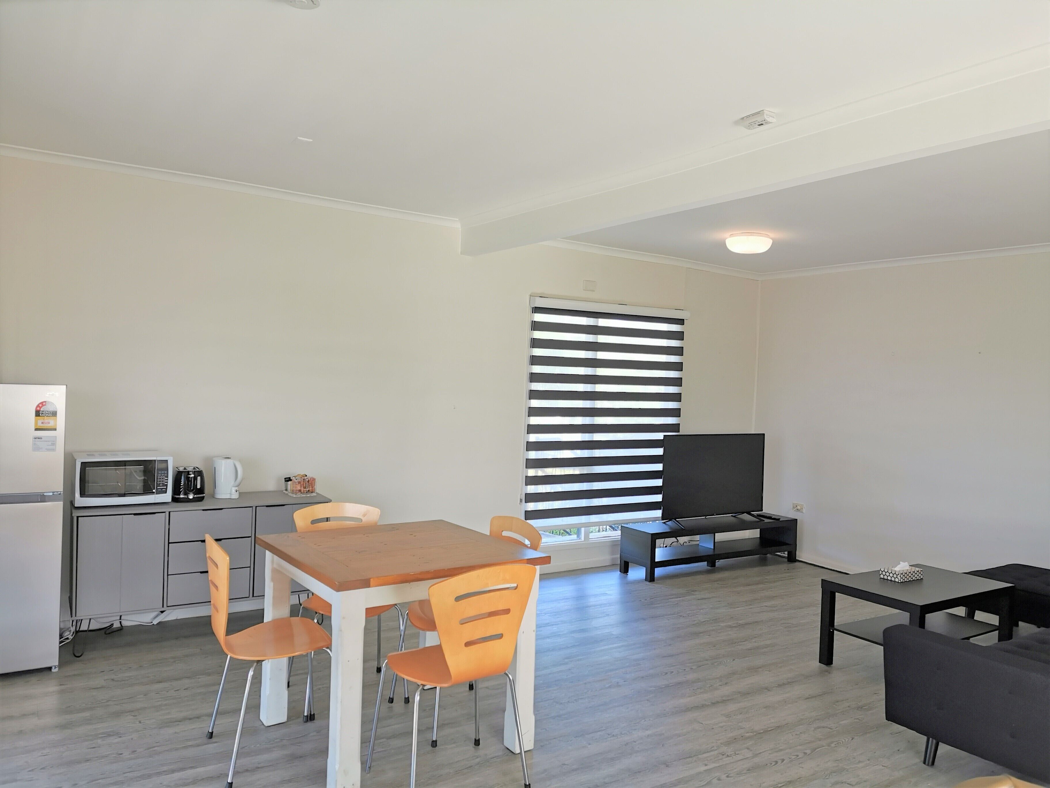 A compact living space with a kitchenette, a dining area, a seating area with a sofa and a TV, and a large window with blinds.