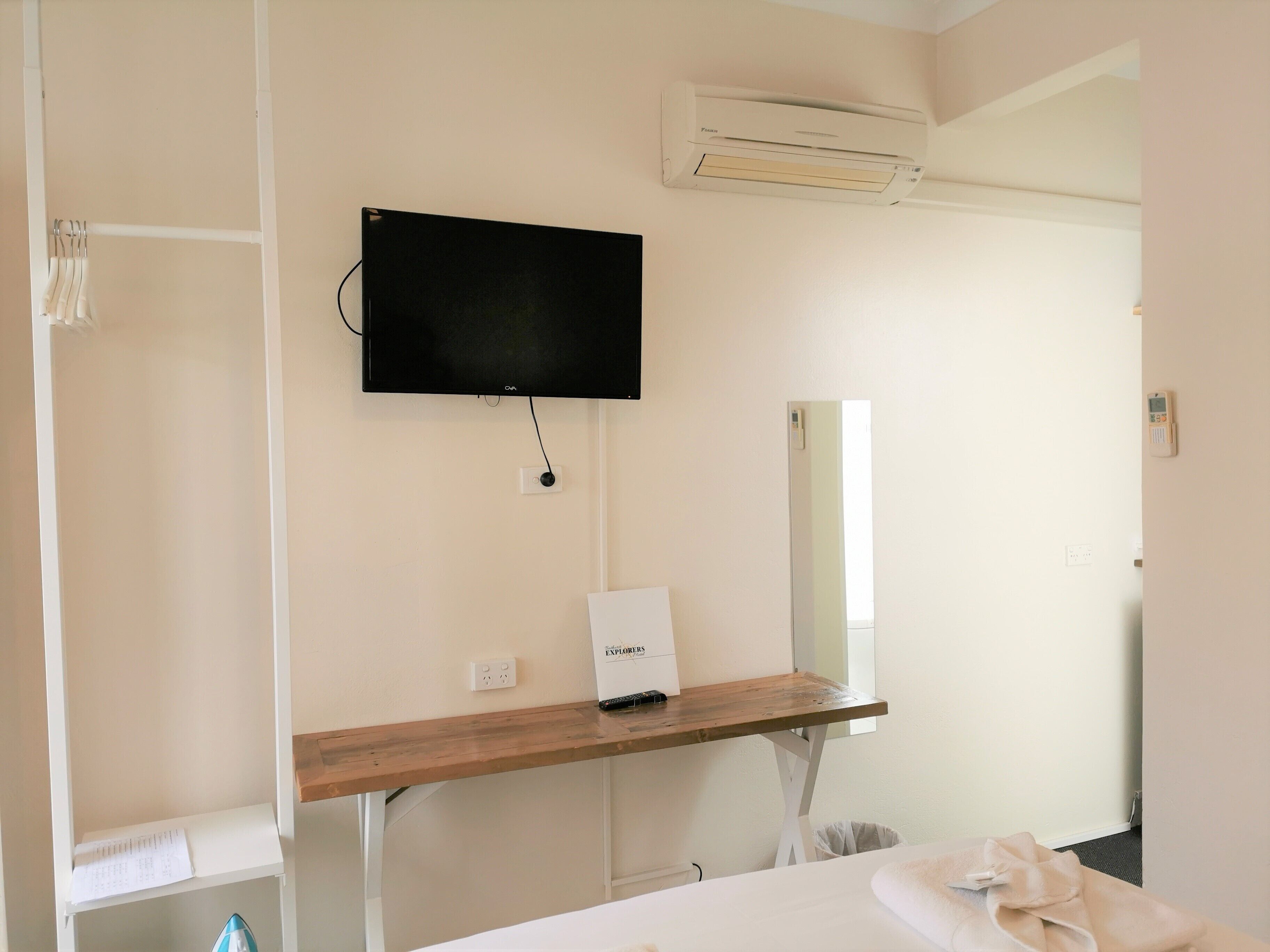 A hotel room with a flat-screen TV mounted on the wall, a wooden desk with a small box labeled "EXPLORERS," a mirror, and a bed with a white comforter.