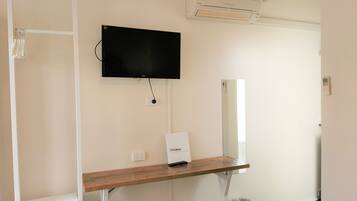 A hotel room with a flat-screen TV mounted on the wall, a wooden desk with a small box labeled "EXPLORERS," a mirror, and a bed with a white comforter.