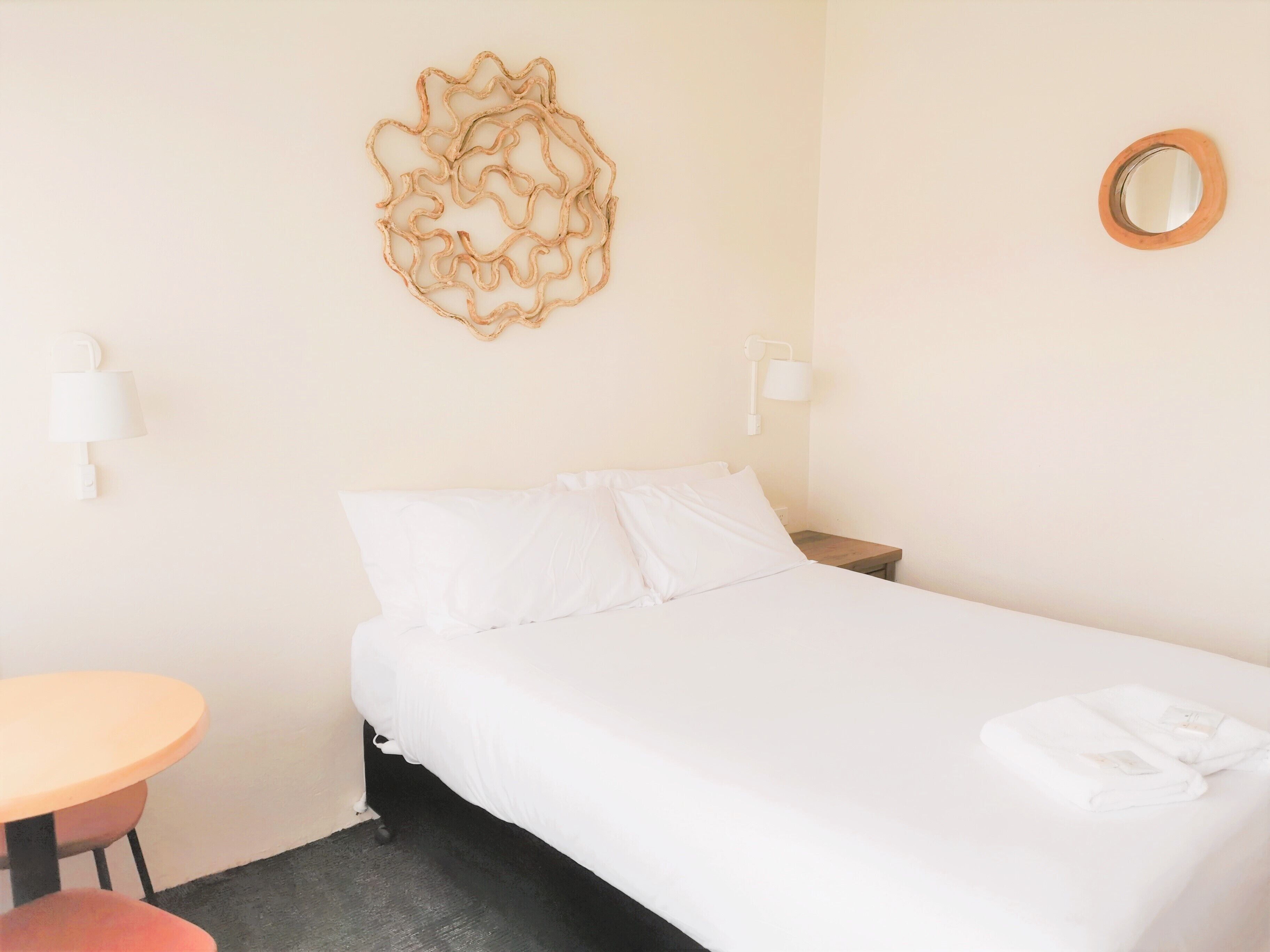 A hotel room with a bed, a round mirror, a wall-mounted lamp, and a small table with a chair.