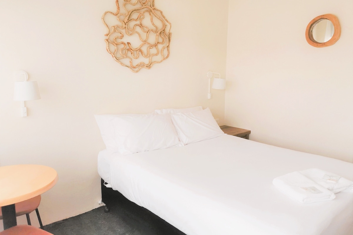 A hotel room with a bed, a round mirror, a wall-mounted lamp, and a small table with a chair.