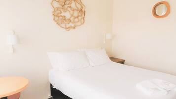 A hotel room with a bed, a round mirror, a wall-mounted lamp, and a small table with a chair.
