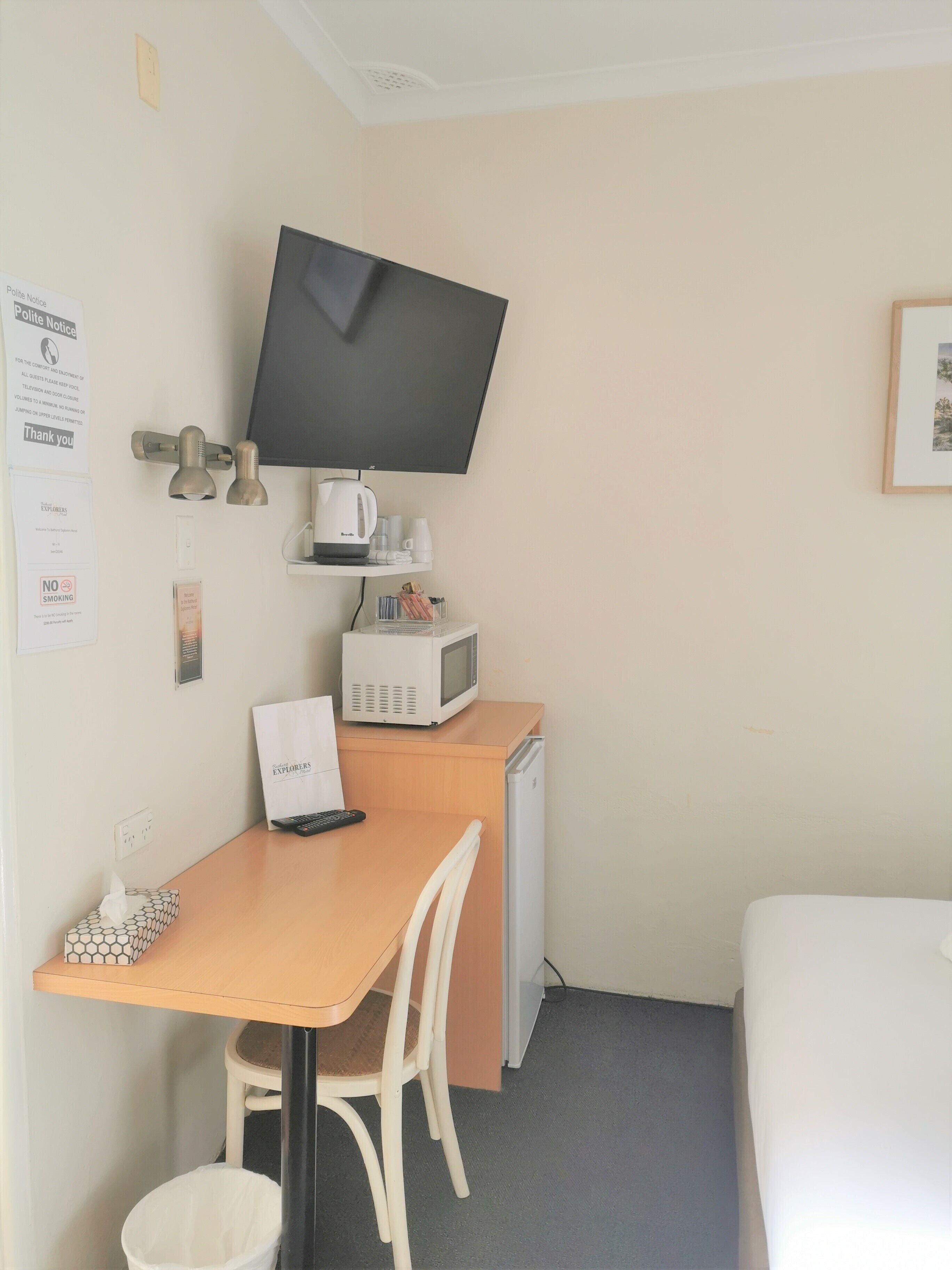 A compact hotel room with a single bed, a small desk with a chair, a television mounted on the wall, a microwave, and a kettle on a shelf.