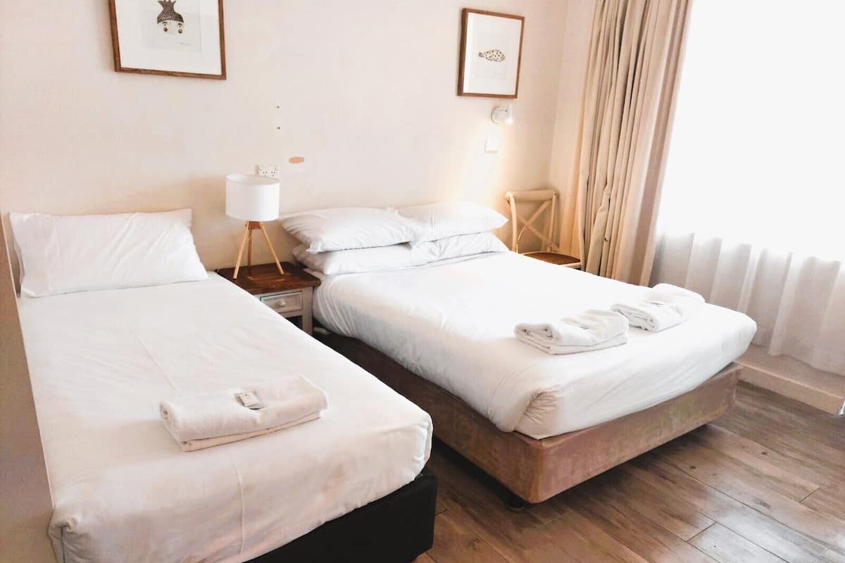 A hotel room with two beds, a bedside table with a lamp, a chair, and a radiator.
