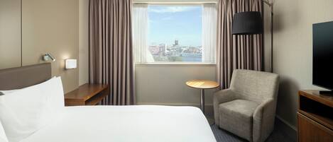 A hotel room with a large bed, a chair, a desk, a TV, and a window with a city view.