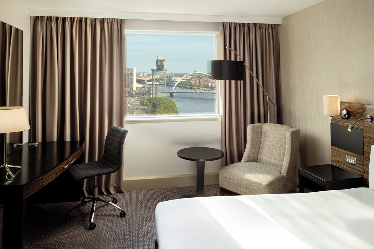 Hotelkamer met een bed, bureau, stoel, klein tafeltje en een lamp. Er is een raam met uitzicht op een brug en gebouwen.