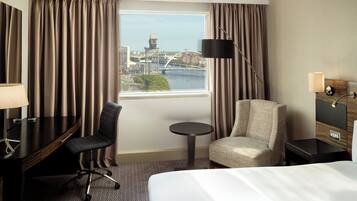 Ein Hotelzimmer mit einem Bett, einem Schreibtisch, einem Stuhl, einem kleinen Tisch und einer Lampe. Ein Fenster bietet Blick auf eine Brücke und Gebäude.