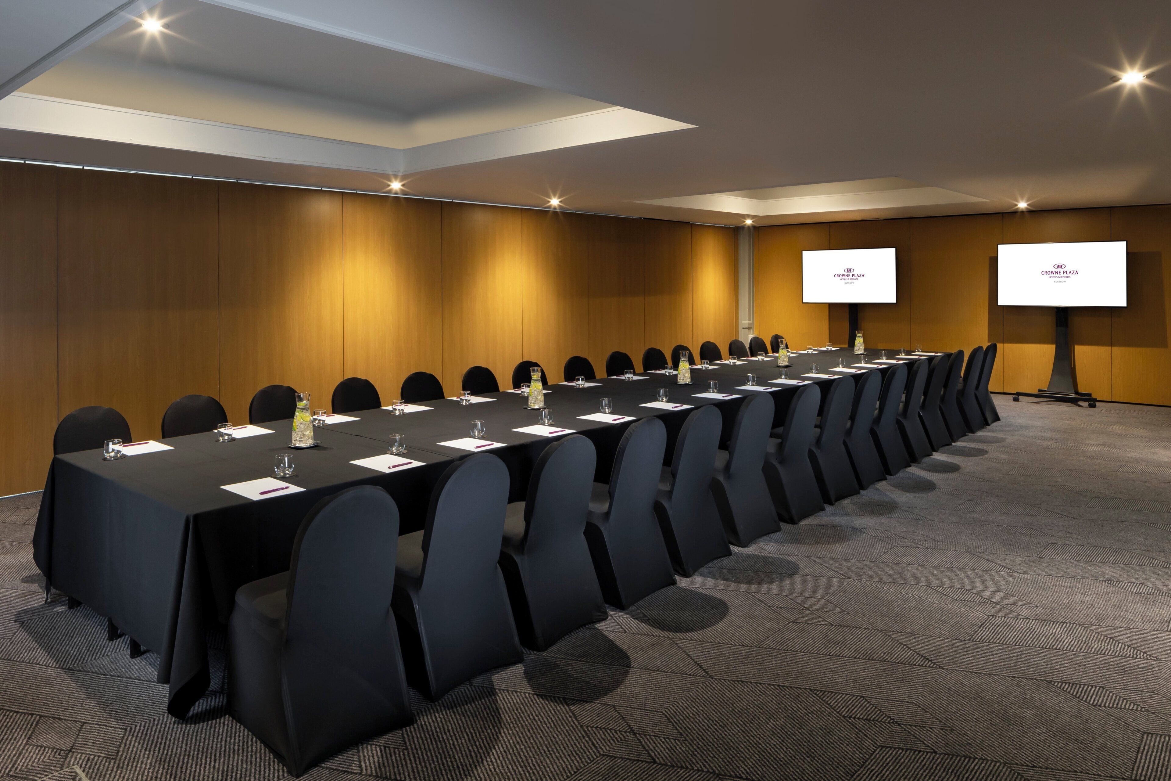 A conference room with a long table, black chairs, and two screens on stands.