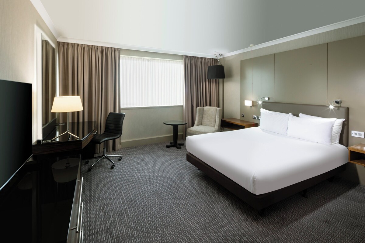 Een hotelkamer met een groot bed, een bureau met lamp, een stoel, een klein tafeltje en een televisie.