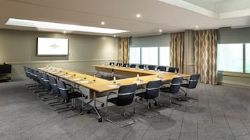 A conference room with a long table, chairs, a projector screen, and a large window with curtains.