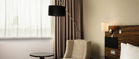 Ein Hotelzimmer mit Bett, Stuhl, kleinem Tisch, Lampe und einem Fenster mit Vorhängen.