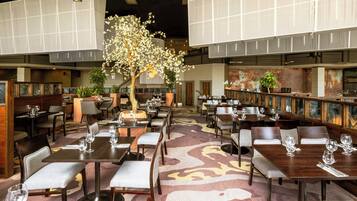 A spacious restaurant with multiple dining tables and chairs, a central decorative tree, and a patterned carpet.