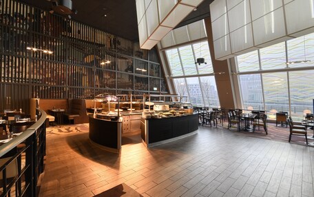 A modern restaurant with a central bar, high ceilings, and large windows offering city views.
