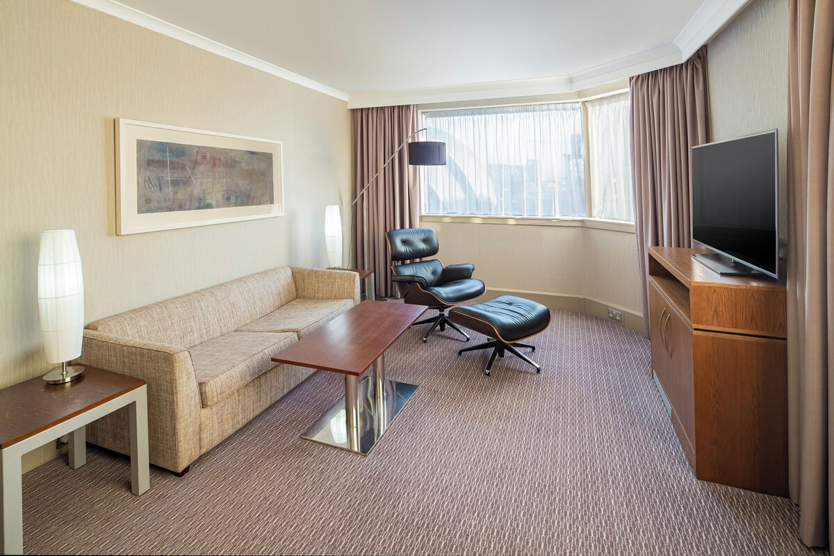 Een hotelkamer met een bank, een tafeltje, een stoel, een televisie en een raam met gordijnen.