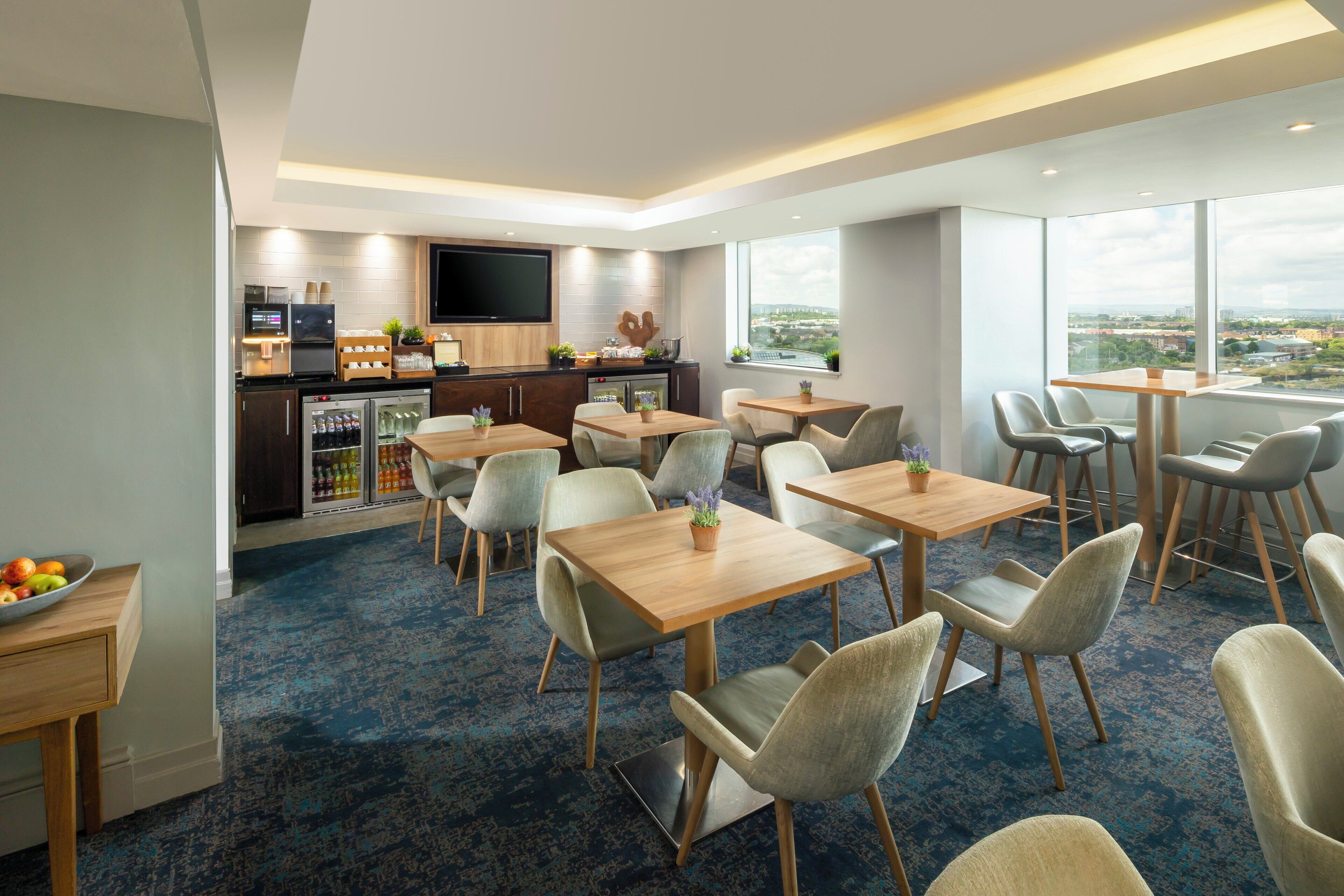 A modern hotel room with a dining area, kitchen, and large windows offering a city view.