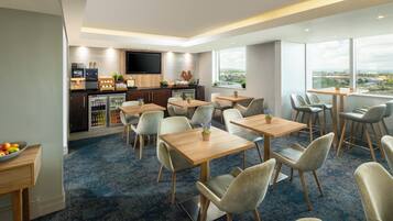 A modern hotel room with a dining area, kitchen, and large windows offering a city view.