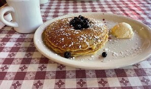 Un plato de panqueques con arándanos y azúcar glas, una bola de helado y una taza blanca sobre un mantel a cuadros.