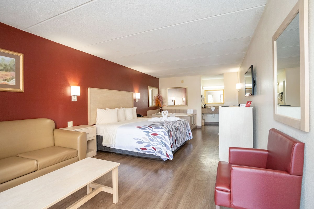 A hotel room with a bed, a red armchair, a beige sofa, a small table, and a mirror.
