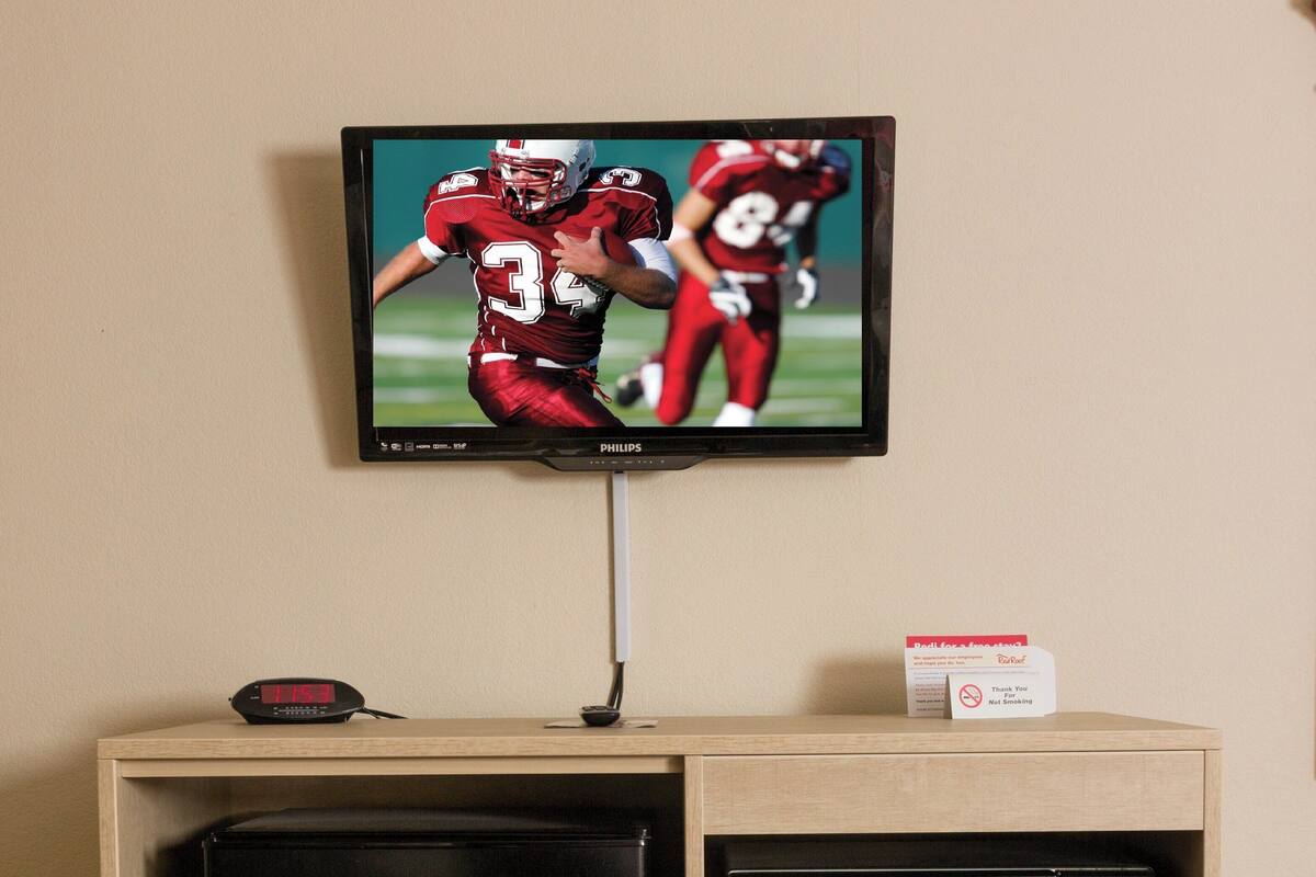 A wall-mounted television displaying a football game, a black refrigerator, and a microwave on a wooden cabinet.