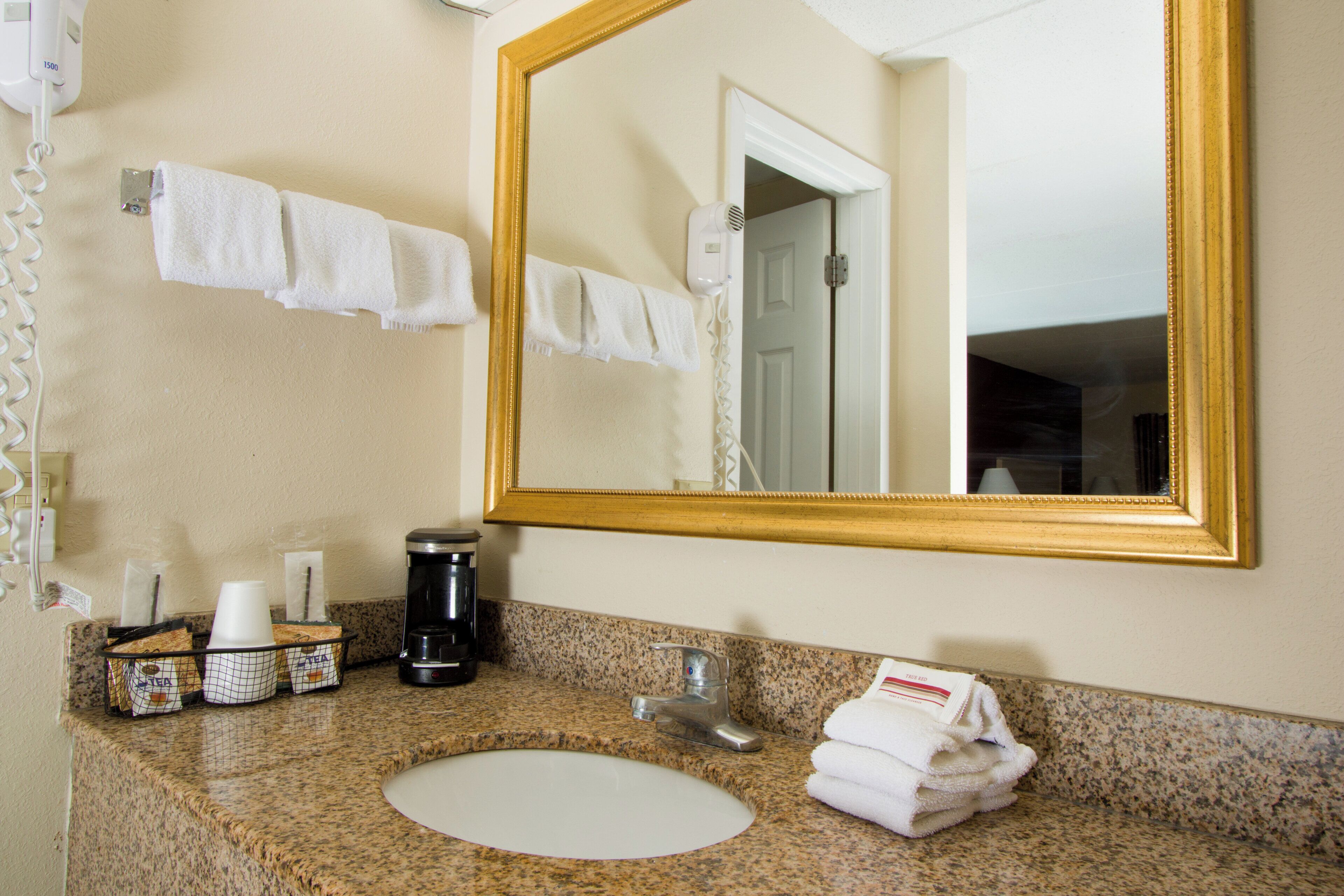 A bathroom with a granite countertop, a white sink, a coffee maker, and a mirror with a gold frame.