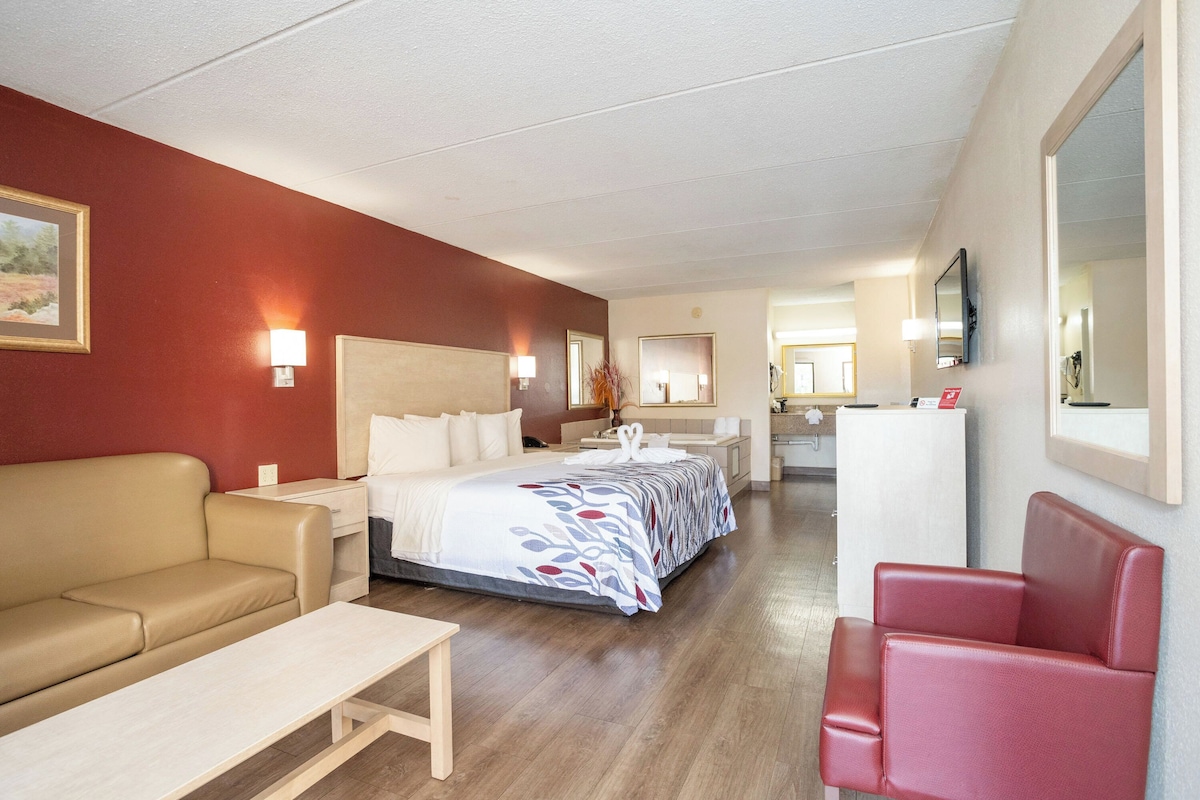 Una habitación de hotel con una cama, un sillón rojo, un sofá color beige, una mesita y un espejo.