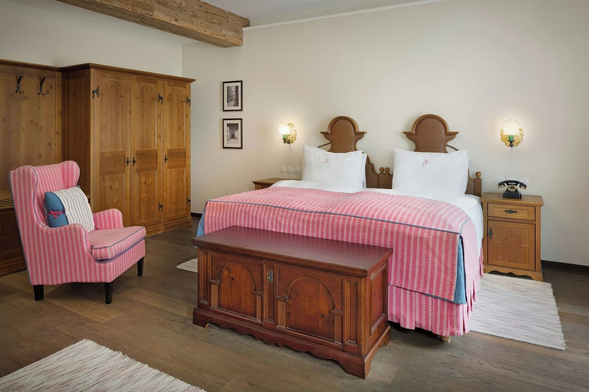 A bedroom with a wooden wardrobe, a pink chair, a bed with a striped bedspread, and two wall-mounted lamps.