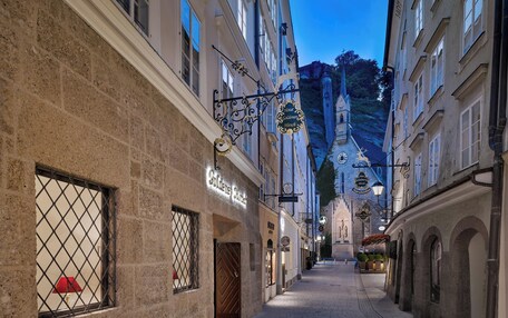 A narrow street with historic buildings, a church spire in the background, and a sign for 'Goldener Hirsch' hotel.