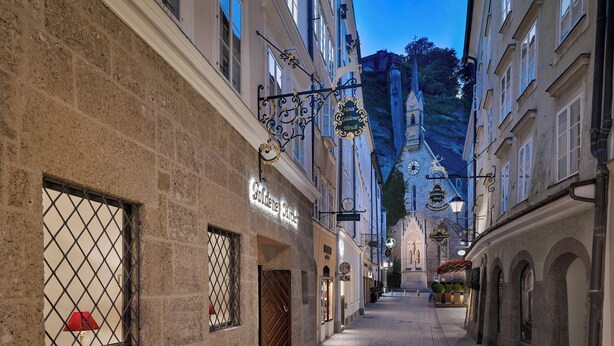 A narrow street with historic buildings, a church spire in the background, and a sign for 'Goldener Hirsch' hotel.