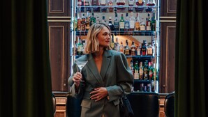 A person in a suit standing in front of a bar with shelves of liquor bottles.