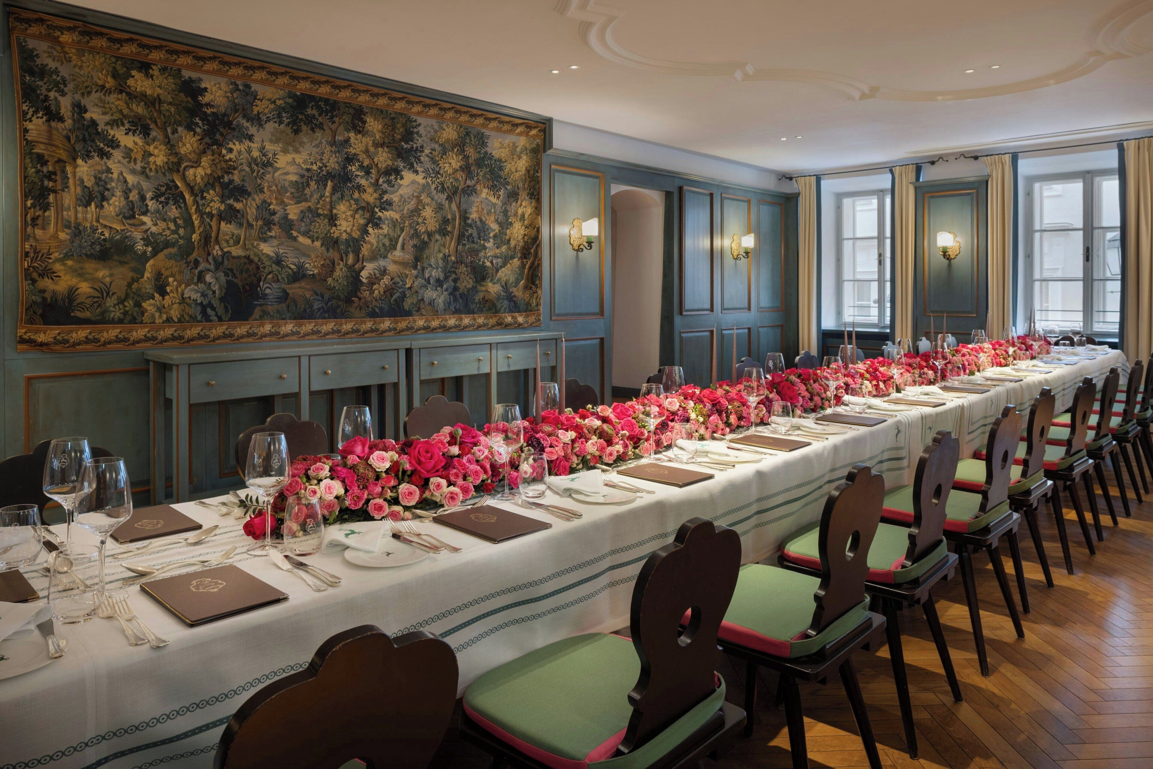 A long dining table set for a formal meal with chairs, a floral centerpiece, and a large painting on the wall.