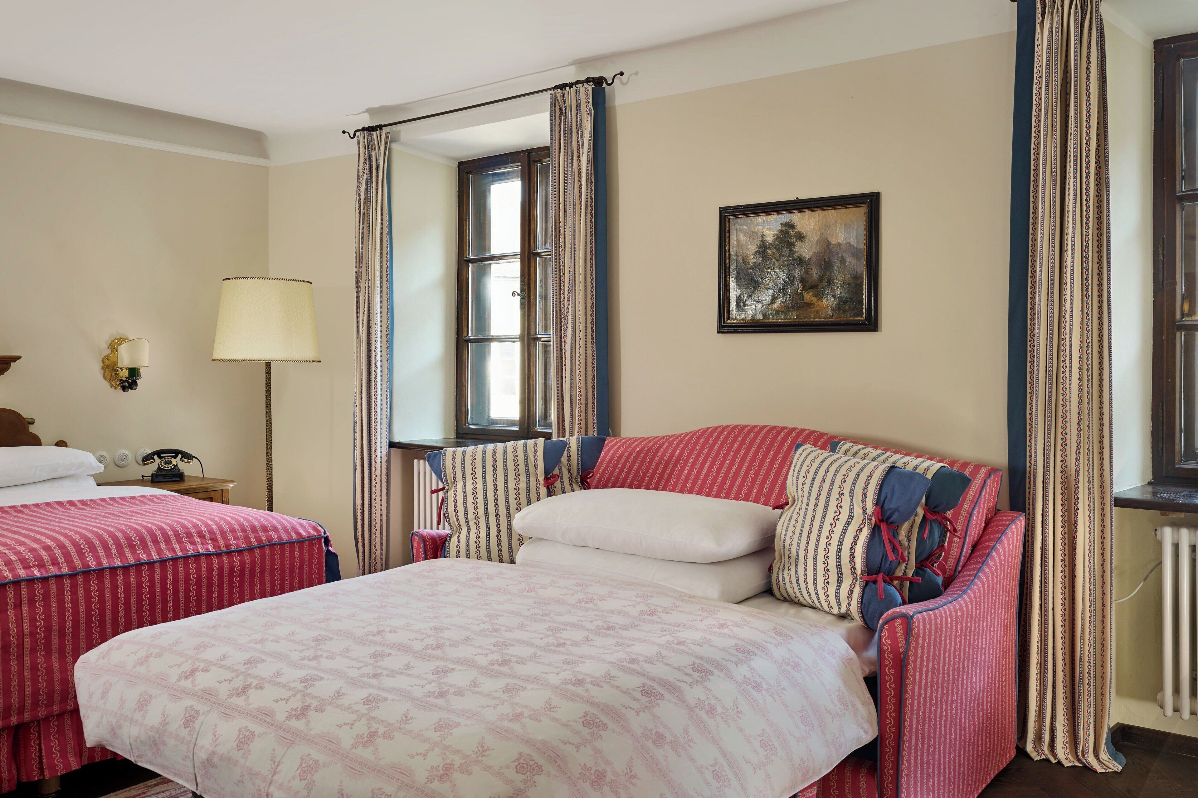 A hotel room with two beds, a bedside table with a lamp, a painting on the wall, and a window with curtains.