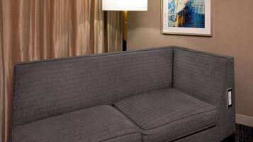 A gray sofa in a hotel room with a lamp and a framed abstract painting on the wall.