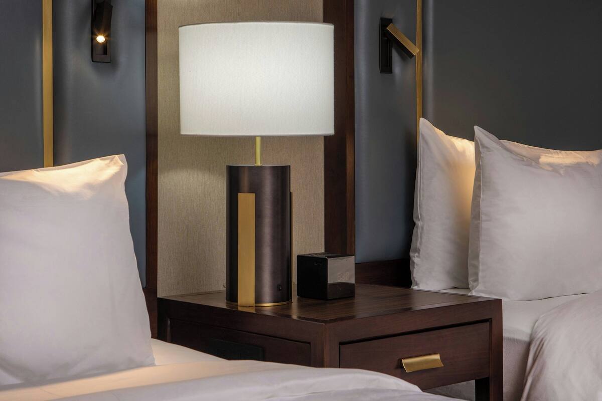 A hotel room with a bed, a nightstand with a lamp, a clock, and a wall-mounted light.