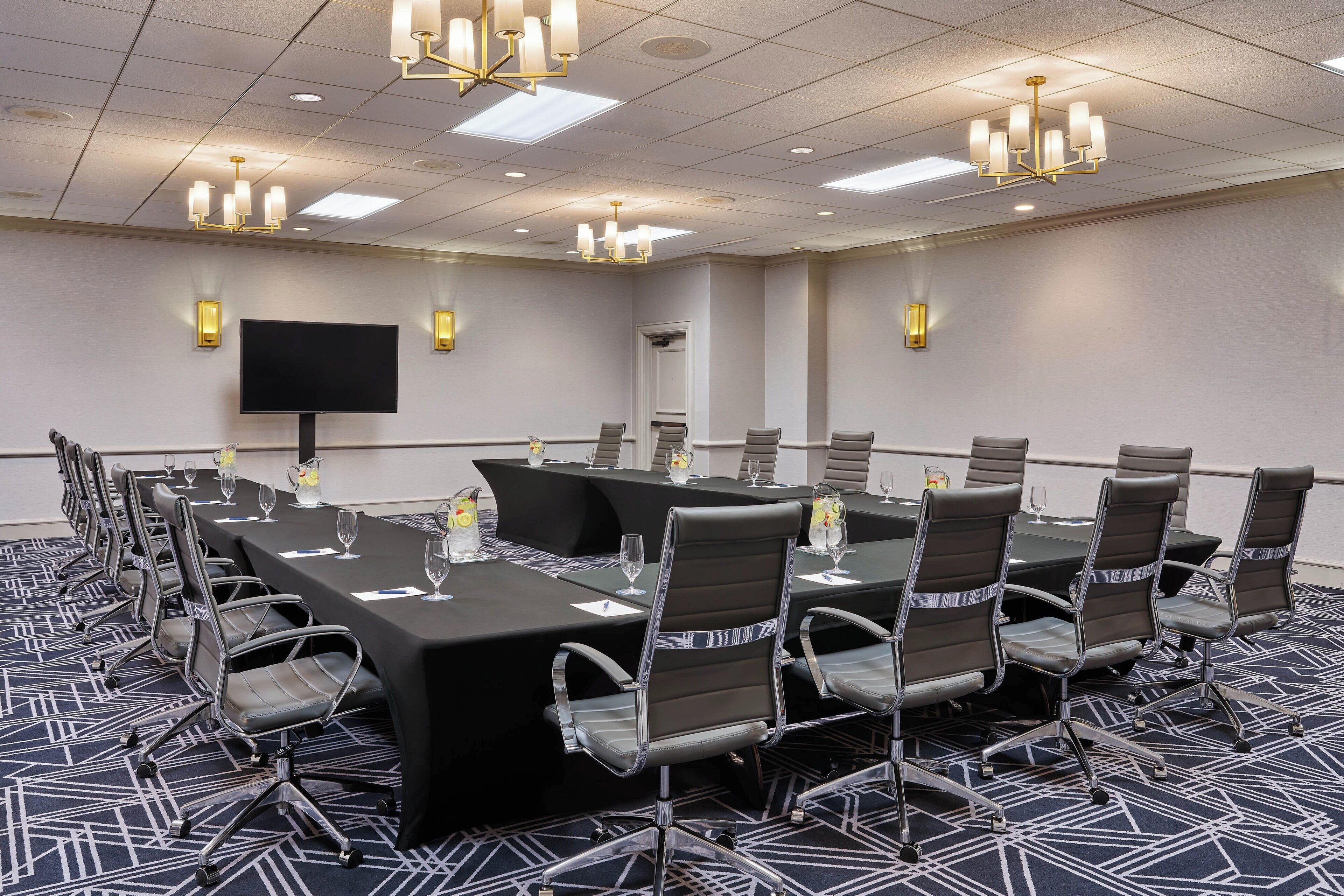 A conference room with a long table, chairs, a flat-screen TV, and chandeliers.