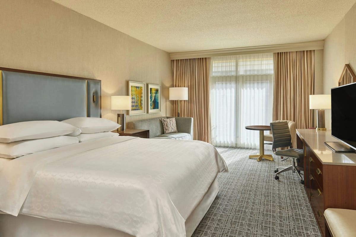 A hotel room with a large bed, a desk with a TV, a chair, a lamp, and a window with curtains.