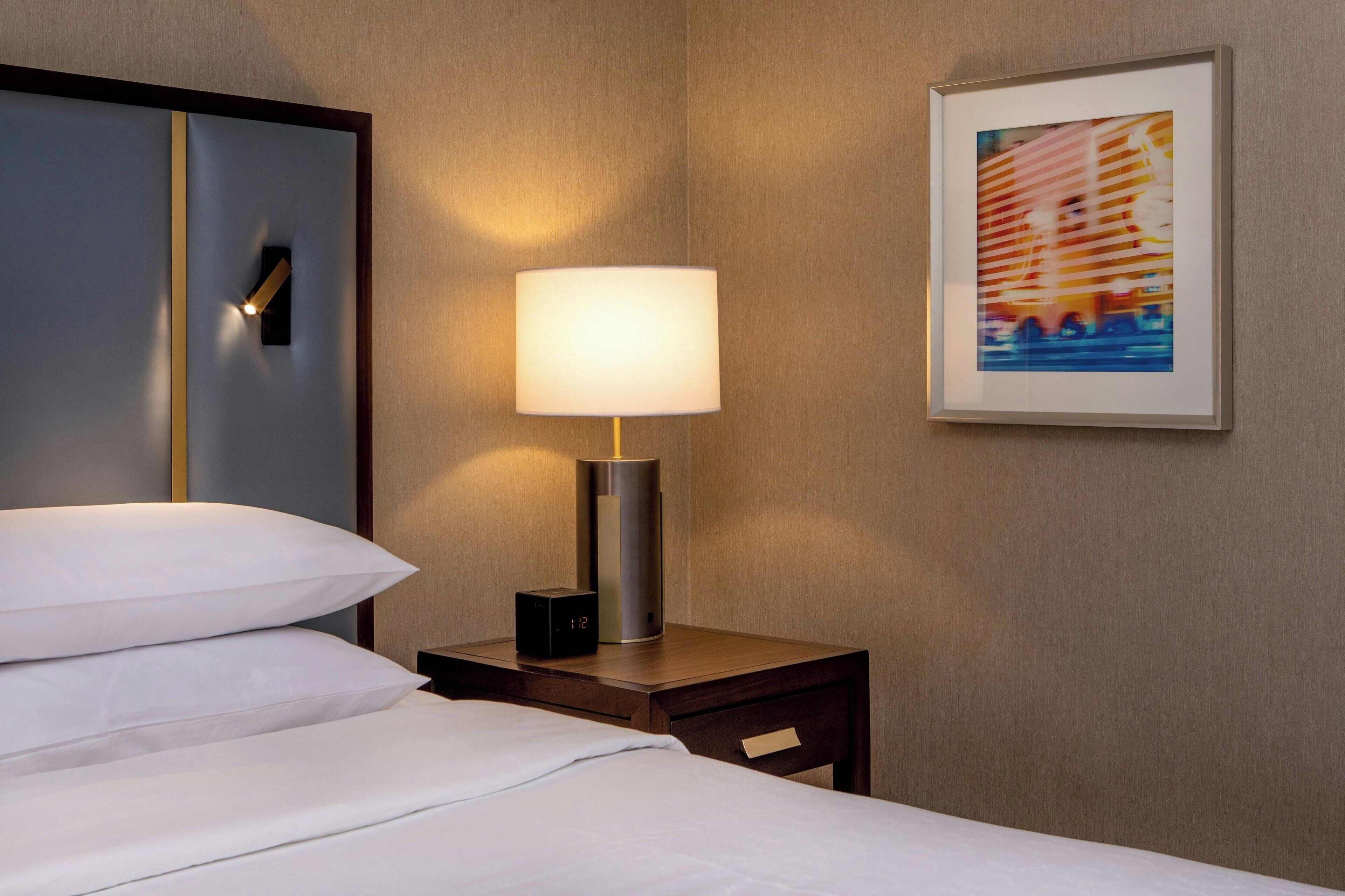 Habitación de hotel con una cama, una mesita de noche con lámpara, un reloj y un cuadro en la pared.