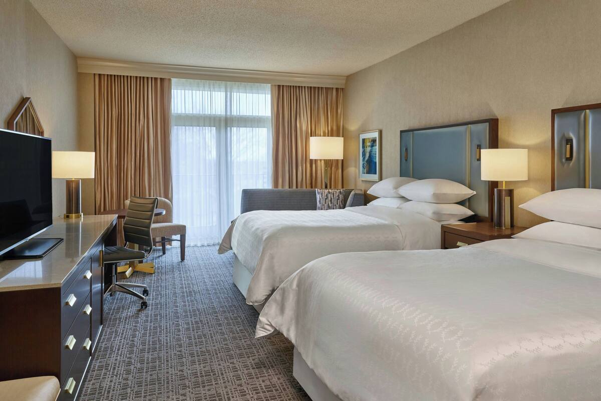 A hotel room with two beds, a desk with a television, a chair, and a large window with curtains.