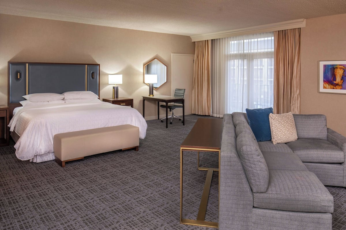 A hotel room with a bed, a desk, a sofa, and a large window with curtains.