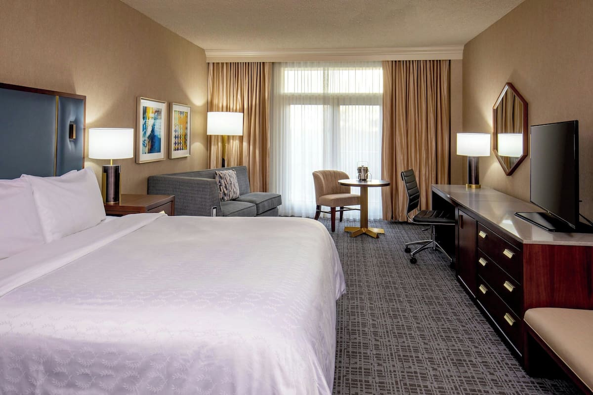A hotel room with a large bed, a desk, a chair, a television, and a small table with a lamp.